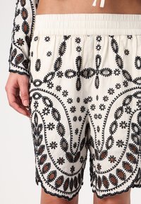 White embroidered shorts featuring black floral designs, textured trim, and an elastic waistband, showcasing intricate detailing and cutouts.