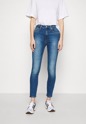 Person wearing slim-fit blue jeans, white oversized t-shirt, and white athletic shoes standing against a plain background.