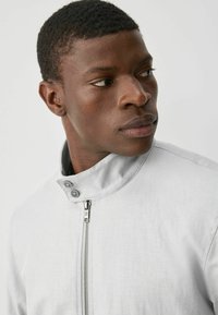 Chaqueta gris con cremallera y cuello alzado, que presenta dos botones y una tela texturizada, mostrada en la parte superior del torso de un modelo.