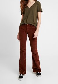 Olivově zelené tričko s výstřihem do V v kombinaci s hnědými korduroidovými kalhotami s rozšířenými nohavicemi. Černé tenisky doplňují celý outfit. Jednoduchý, ležérní design.