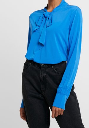 Kobieta w jasnoniebieskiej bluzce z długim rękawem, zawiązanej na kokardę przy szyi, oraz czarnych, wysokowcześnie dopasowanych dżinsach na tle jednolitego tła.