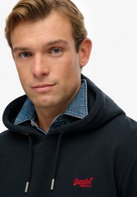 Felpa navy con logo rosso "SuperDry", caratterizzata da cappuccio con cordini e colletto di camicia in denim che spunta. Tessuto liscio, design casual.