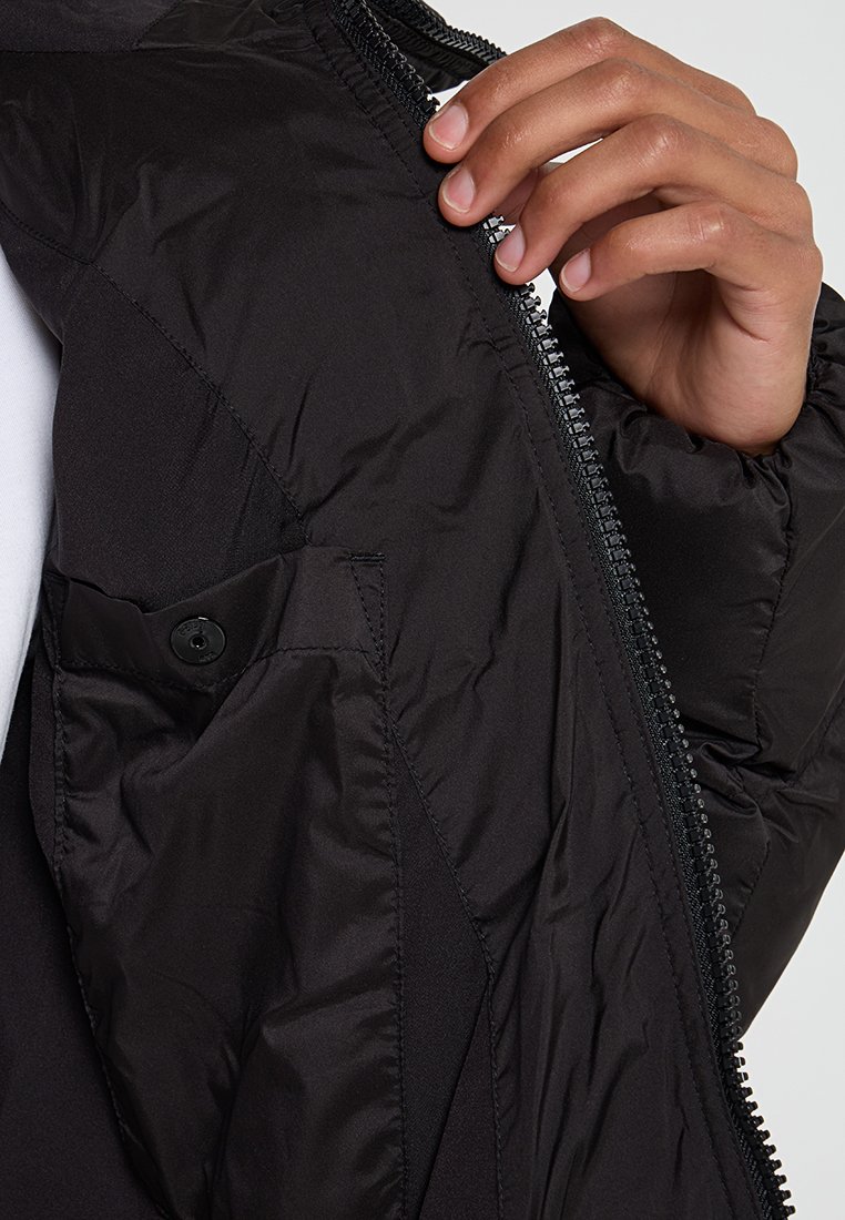 Schwarze gepolsterte Jacke mit einer glatten, matten Textur. Verfügt über einen Reißverschluss und eine mit Knopfverschluss versehene Tasche auf der linken Seite, die teilweise geöffnet dargestellt wird.