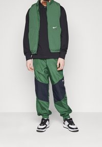 Man som bär gröna och svarta Nike-trackbyxor, grön väst, svart långärmad tröja och svart-vita Nike-sneakers, står mot en enfärgad bakgrund.