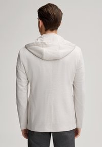 Giacca con cappuccio grigio chiaro, texture morbida, cuciture minime e due tasche laterali, vestibilità aderente, indossata sopra pantaloni scuri.