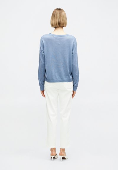 Γυναίκα με κοντά ξανθά μαλλιά, που φοράει ανοιχτό μπλε πουλόβερ, λευκό παντελόνι ίσια γραμμή και λευκά ψηλοτάκουνα παπούτσια, στέκεται με την πλάτη στραμμένη.