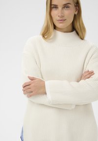 Femme avec de longs cheveux blonds portant un pull en col roulé blanc à côtes, les bras croisés, sur un fond clair uni.