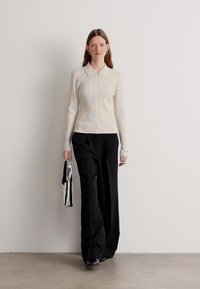 Lys kremfarget strikket skjorte med krage, kombinert med vide svarte bukser og holder en sølvstripet veske. Flate sko fullfører antrekket.