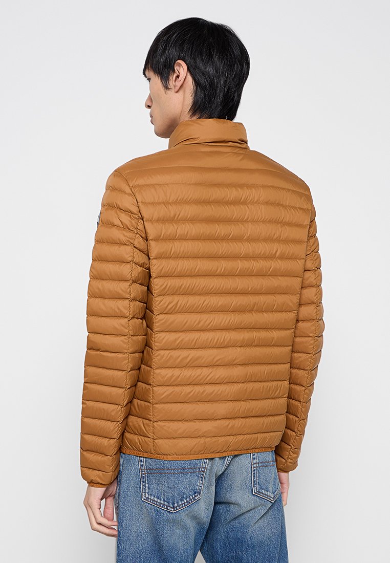 Giacca imbottita marrone con cuciture orizzontali, colletto rialzato e una texture liscia. Indossata sopra dei jeans in denim blu con cuciture delle tasche visibili.