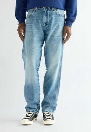 Personne portant un jean droit bleu clair, des baskets noires et blanches à lacets, et un haut bleu à manches longues, debout devant un fond blanc.