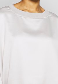 Primer plano de una persona que lleva una blusa de satén gris claro, suave, con un escote ancho y acanalado.