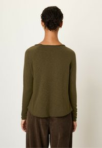 Long-sleeve olive green top with a rounded neckline and raw edges, paired with brown corduroy pants featuring vertical stripes.