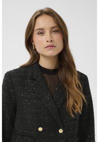 Juodas tekstūruotas švarkas su dvispiegiu dizainu, puoštas auksiniu atspalviu mygtukais ir po juo aukštos kokybės permatomu juodu megztiniu. Lygi plaukų tekstūra su subtiliomis bangomis.