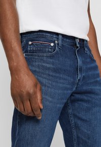 Vaqueros de denim azul oscuro con corte recto, que cuentan con un acento rojo en el bolsillo delantero y herrajes plateados. Textura suave, lavado medio.
