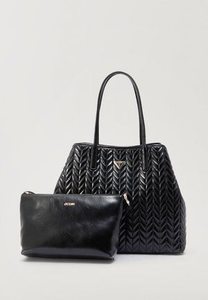 Sac cabas noir matelassé avec longues poignées et petite pochette noire zippée, tous deux ornés de logos Guess dorés, sur fond gris.