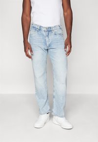 Jeans de mezclilla azul claro con un corte relajado, que presentan desvanecidos y sutiles detalles desgastados. Combinados con zapatillas blancas y una camiseta sin mangas blanca.