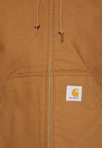 Brown, zip-up hoodie made of durable fabric with a kangaroo pocket, drawstring hood, and a square logo patch in the lower right corner.