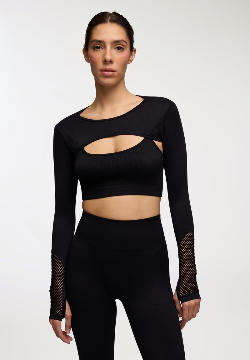 Crop top nero a maniche lunghe con design a taglio, texture a coste e dettagli in rete sulle maniche. Abbinato a leggings neri a vita alta.