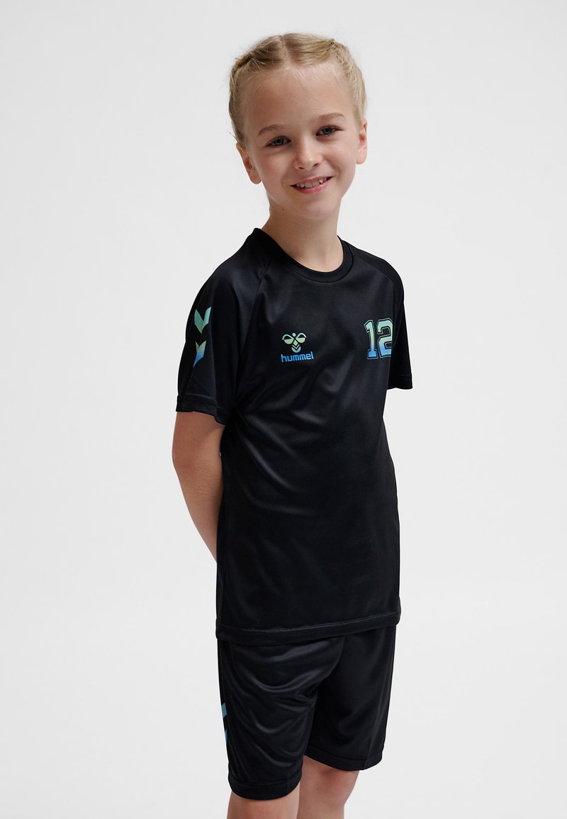 Lachend kind met gevlochten haar, gekleed in een zwart sportshirt en short met blauwe nummer 12 en hummel-logo's, staand met de handen achter de rug.