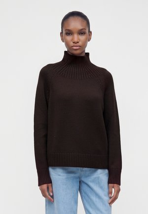 Kvinde iført en mørkebrun ribstrikket rullekravetrøje og lyseblå jeans, der står mod en ensfarvet lys baggrund.