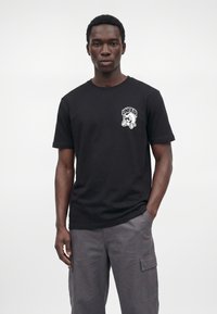 Mann trägt ein schwarzes T-Shirt mit weißem "UNFAIR" Cartoon-Logo auf der Brust und graue Cargohosen, steht vor einem weißen Hintergrund.