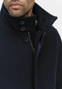 Marine uld frakke med høj krave, udstyret med en lynlås og fire sorte knapper. Glat tekstur med en struktureret design.