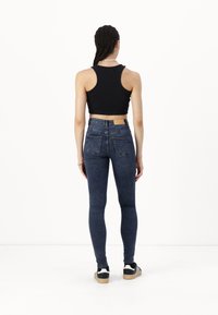 Black cropped tank top and blue skinny jeans, back view; jeans feature two back pockets and a standard waistband. Footwear is black and white sneakers.