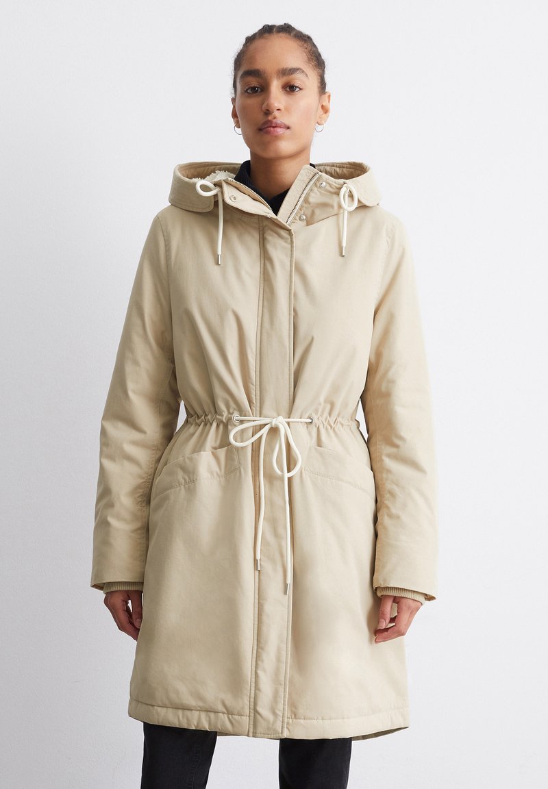 Beige Parka mit Kapuze, Kordelzug in der Taille, langen Ärmeln, zwei Fronttaschen und einem Reißverschluss mit Druckknopfverschluss. Weicher, strukturierter Stoff.