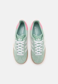 Zelené semišové tenisky s bílými pruhy, růžovými akcenty a bílými tkaničkami. Mají texturovanou podrážku pro lepší přilnavost a vnitřek s brandingem.