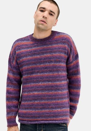 Jeune homme portant un pull en tricot rayé violet et orange, debout devant un fond clair uni.