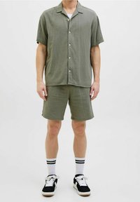 Olijfgroene shirt met korte mouwen en een knoopsluiting aan de voorkant, gecombineerd met bijpassende shorts. Heeft een gestructureerde stof en wordt gedragen met zwarte schoenen en gestreepte sokken.