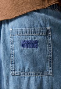 Spodnie dżinsowe z naszywaną kieszenią ozdobioną niebieskim haftem "American Vintage". Materiał ma jasne wykończenie, a faktura jest lekko sprana.