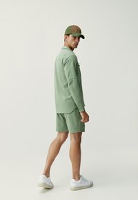 Ljusgrön kortärmad skjorta och matchande shorts, tillverkade av glatt tyg. Brun keps med nätbak. Vita sneakers.