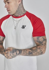Tricou raglan cu mâneci scurte, roșu și alb, având un logo brodat negru pe piept și o textură de bumbac neted.