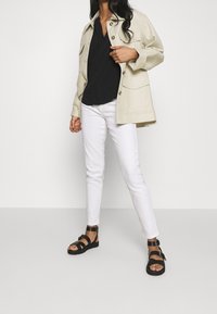 Chaqueta beige clara con botones, blusa negra de escote en v, jeans blancos ajustados y sandalias negras con tiras y detalles de hebilla. Texturas suaves en todo.