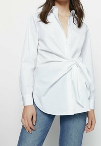 Chemise blanche à boutons avec manches longues, un détail de nœud à l'avant et un ourlet arrondi. Le tissu semble lisse et léger.