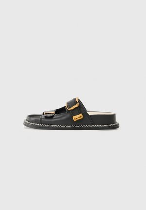 Sorte skinnslide-sandaler med to justerbare stropper med gullfargede spenner, en flat såle og hvit søm langs kantene.