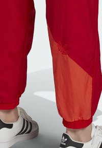 Piros széldzseki nadrág átlós narancssárga panellel, rugalmas mandzsettákkal és dombornyomott Adidas logóval. Fehér sportcipőkkel viselve, fekete csíkokkal.