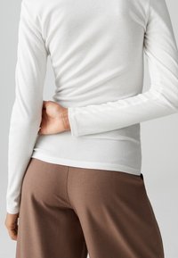 Person trägt ein weißes langärmeliges Hemd und braune Hose, mit einer Hand, die auf dem unteren Rücken ruht, vom Betrachter weg gewandt.