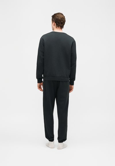 Άνδρας με την πλάτη γυρισμένη, φοράει μαύρο φούτερ και μαύρη φόρμα, στέκεται σε λευκό φόντο.