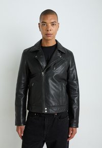 Mustast nahast mootorratturi jope, millel on nurgeline krae, mitte-sümmeetriline tõmblukku sulgemine ja küljetaskud; tekstureeritud pind ja hõbedased detailid.