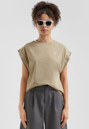 T-shirt en coton beige avec des manches retroussées, coupe décontractée, et petit logo blanc sur la poitrine, associé à un pantalon large gris foncé.