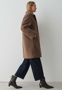 Manteau en teddy marron avec un matériau texturé et duveteux, comportant un col et des poches. Associé à un pantalon large bleu foncé et des bottines noires.