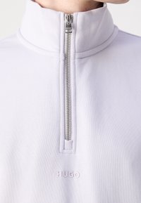 Sweat-shirt blanc HUGO à demi-fermeture éclair argentée avec un col montant porté autour du cou et de la partie supérieure de la poitrine d'une personne.