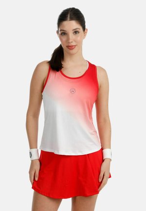 Junge Frau mit dunklem Haar im Pferdeschwanz, trägt ein rot-weißes, verlaufendes ärmelloses Oberteil, roten Rock und weiße Handgelenkbandagen, steht vor neutralem Hintergrund.