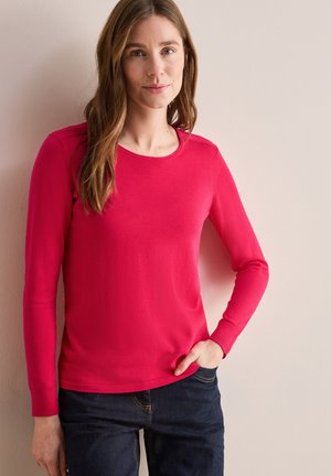 Maglietta aderente a maniche lunghe in cotone rosa brillante, con scollo tondo e polsini a costine, abbinata a jeans scuri. Texture liscia.