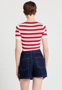 Person mit kurzen braunen Haaren, die ein rot-weiß gestreiftes Kurzarmoberteil und dunkelblaue Jeansshorts trägt, von hinten gesehen.