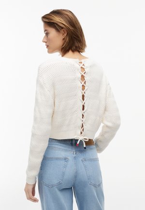 Kvinde med kort brunt hår iført en hvid strikket trøje med åbent rygspil og lyseblå højtaljede jeans, vendt mod venstre.