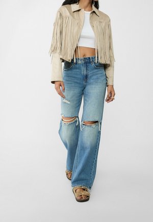 Femme portant une veste à franges beige, un top court blanc, un jean bleu large déchiré et des sandales beige marchant sur un fond uni.