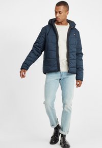 Navy padded jacket with a hood, front zip closure, and side pockets. Worn over a light gray sweater and paired with light blue jeans.